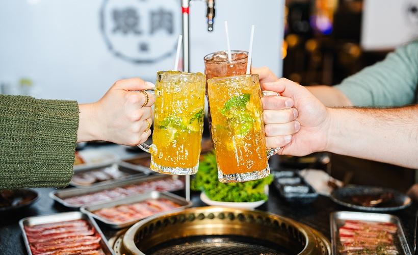 Two people toasting tall glasses of iced tea over a Korean BBQ grill